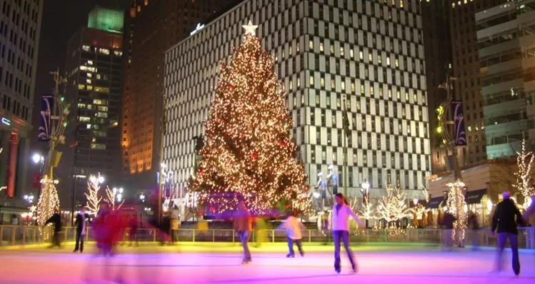 Skaters on an illuminated outdoor rink beside a lit holiday tree in a city center
